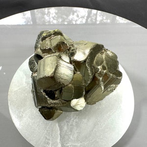 Könnte beinhalten: Ein großer, metallischer, goldfarbener Pyritkristall-Cluster. Der Kristall besteht aus vielen einzelnen, facettierten Kristallen, die zusammen gruppiert sind. Der Kristall liegt auf einer weißen, runden Oberfläche.