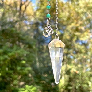 Puede incluir: Un péndulo de cristal de cuarzo transparente con una cadena de plata y un dije de plata Om. El péndulo cuelga sobre un fondo borroso de follaje verde.