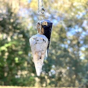 May include: Two crystal pendulums, one clear with white flecks and one black, hanging from silver chains against a blurred green background.