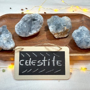 Puede incluir: Cuatro grupos de cristales de celestita azul claro en una bandeja de madera con un pequeño letrero de pizarra que dice "celestita".