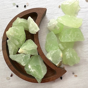 May include: A collection of green celestite crystal chunks in a wooden crescent moon-shaped dish. The crystals are various sizes and shapes, with some having a translucent quality.
