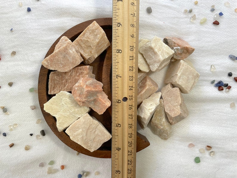 May include: A collection of pink and white selenite crystals in a wooden crescent moon-shaped dish. The crystals are various sizes and shapes. A ruler is next to the dish for scale.