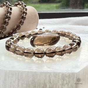 Puede incluir: Una pulsera de cuarzo ahumado con una piedra grande de forma ovalada en el centro. La pulsera está hecha de cuentas redondas y translúcidas. Una segunda hebra de cuentas es visible en el fondo. Los artículos se exhiben sobre una superficie blanca.