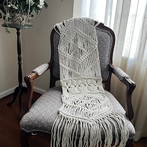 May include: Cream-colored macrame table runner with geometric patterns and fringe. The handmade textile is draped over a vintage upholstered chair with dark wood trim. A decorative plant stand and window are in the background.