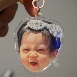 May include: A clear plastic keychain with a photo of a baby girl wearing a blue and white checkered bow. The baby is smiling and looking to the side.