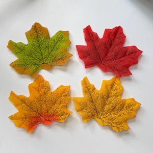 May include: Four artificial maple leaves arranged in a grid. One leaf is green and yellow, one is bright red, one is orange and yellow, and one is solid yellow. Each leaf features a detailed, dark brown printed vein pattern across the surface.