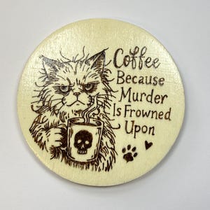 May include: A circular light-colored wooden coaster featuring a wood-burned illustration of a grumpy long-haired cat holding a mug with a skull design. The text reads: Coffee Because Murder Is Frowned Upon, accompanied by a paw print and a heart.