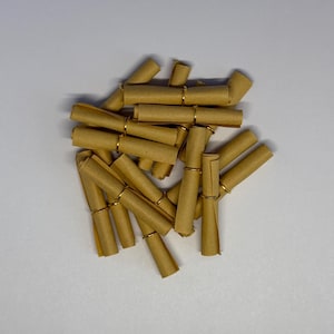 May include: A collection of small, rolled-up scrolls made of tan paper, secured with gold-colored metal rings. The scrolls are scattered across a white surface, creating a textured, layered effect. The paper has a slightly aged appearance.