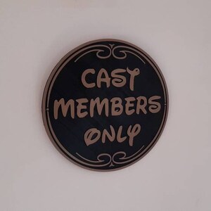 Puede incluir: Un letrero redondo negro con un borde marr&oacute;n y letras doradas que dice "Cast Members Only".