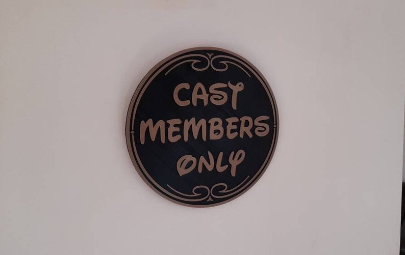 Puede incluir: Un letrero redondo negro con un borde marr&oacute;n y letras doradas que dice "Cast Members Only".