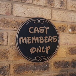 Puede incluir: Un letrero redondo negro y dorado con el texto "CAST MEMBERS ONLY" en una pared de ladrillo.