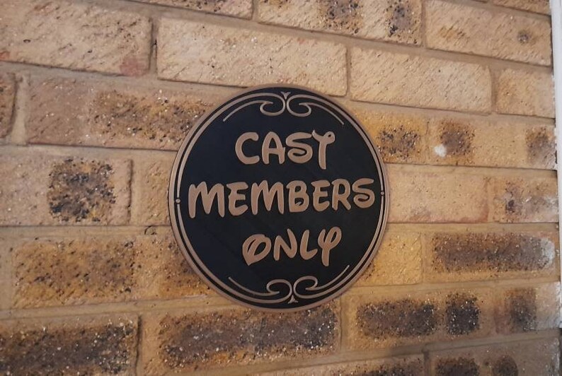 Puede incluir: Un letrero redondo negro y dorado con el texto "CAST MEMBERS ONLY" en una pared de ladrillo.