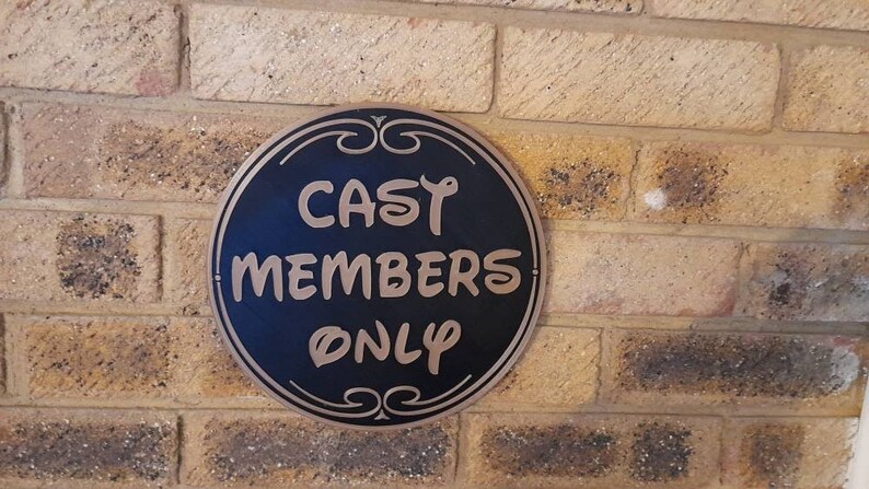 Puede incluir: Un letrero redondo negro con letras doradas en una pared de ladrillo. El letrero dice "Cast Members Only".