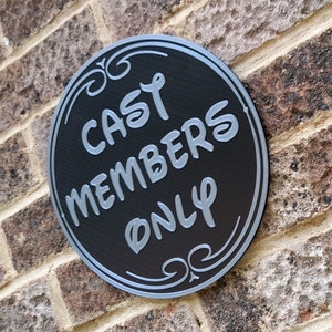 Puede incluir: Un letrero redondo negro con letras plateadas que dice "Cast Members Only" en una pared de ladrillo.