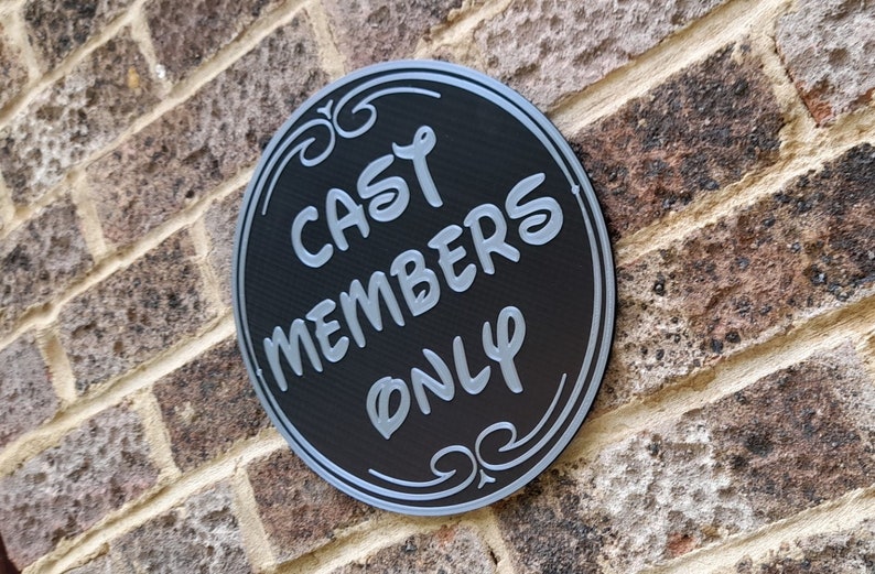 Puede incluir: Un letrero redondo negro con letras plateadas que dice "Cast Members Only" en una pared de ladrillo.