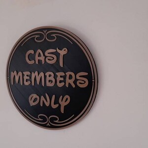Puede incluir: Un letrero redondo negro y dorado con el texto "CAST MEMBERS ONLY" en una fuente decorativa.