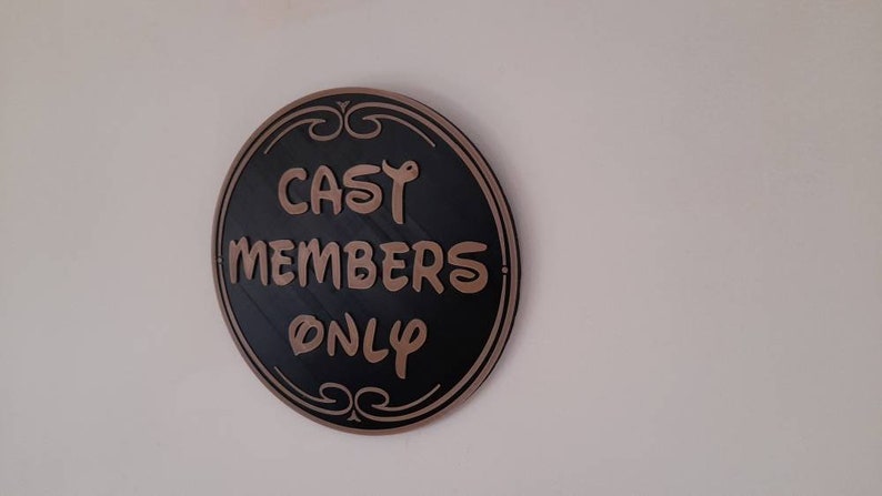 Puede incluir: Un letrero redondo negro y dorado con el texto "CAST MEMBERS ONLY" en una fuente decorativa.