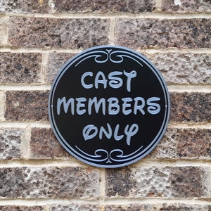 Puede incluir: Un letrero redondo en blanco y negro con el texto "CAST MEMBERS ONLY" en fuente plateada. El letrero est&aacute; montado en una pared de ladrillo.