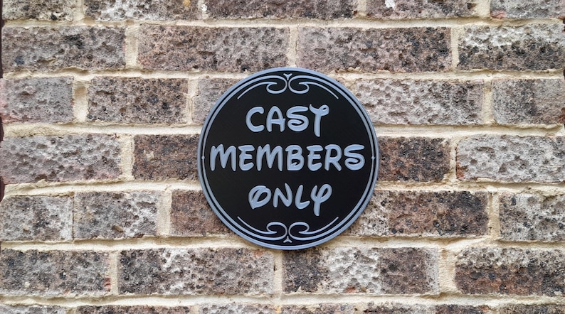 Puede incluir: Un letrero redondo en blanco y negro con el texto "CAST MEMBERS ONLY" en fuente plateada. El letrero est&aacute; montado en una pared de ladrillo.