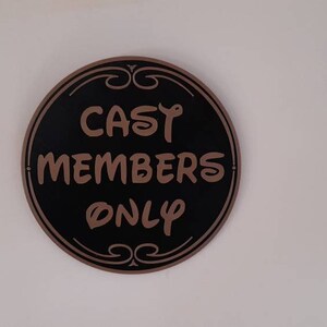 Puede incluir: Un letrero redondo negro y dorado con el texto "CAST MEMBERS ONLY" en letras marrones.