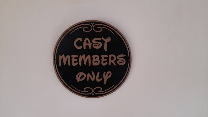 Puede incluir: Un letrero redondo negro y dorado con el texto "CAST MEMBERS ONLY" en letras marrones.
