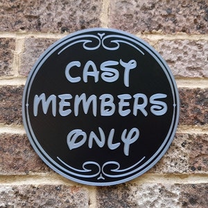 Puede incluir: Un letrero redondo negro y plateado con el texto "CAST MEMBERS ONLY" en blanco.