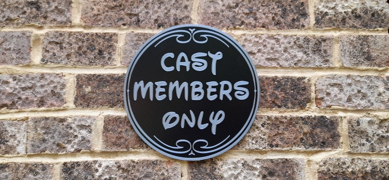 Puede incluir: Un letrero redondo negro y plateado con el texto "CAST MEMBERS ONLY" en blanco.