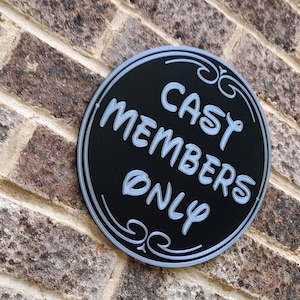 Puede incluir: Un letrero circular negro con un borde blanco y adornos de desplazamiento. El letrero dice "Cast Members Only" en letras blancas.