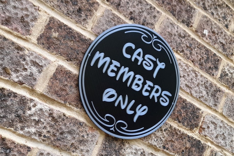Puede incluir: Un letrero circular negro con un borde blanco y adornos de desplazamiento. El letrero dice "Cast Members Only" en letras blancas.