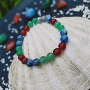 Op de afbeelding: Een armband gemaakt van rode, groene en blauwe edelsteen kralen met een zilveren sluiting. De armband ligt op een witte schelp.