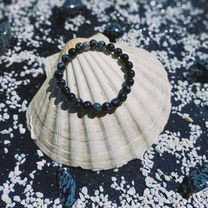 Op de afbeelding: Een armband van zwart-wit gevlekte steen op een witte schelp. De armband is gemaakt van ronde kralen en heeft een zilveren sluiting.