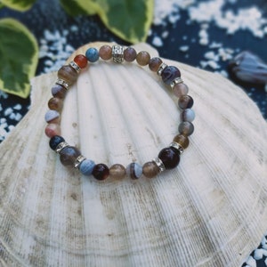 Puede incluir: Un brazalete de ágata marrón, gris y azul con cuentas plateadas y un dije plateado. El brazalete está sobre una concha blanca.