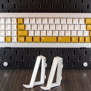 May include: A white keyboard with gold keys and a white frame. The keyboard is mounted on a black pegboard wall. Two white plastic keyboard stands are in the foreground.