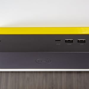 Op de afbeelding: Een zwart Dell laptop dockingstation met een gele bovenkant. Het dockingstation heeft vier poorten aan de achterkant, waaronder een USB-C-poort, twee USB-A-poorten en een stroompoort. Het dockingstation is aangesloten op een stroomkabel.