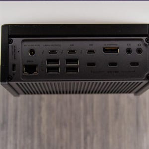 Op de afbeelding: Een zwart elektronisch apparaat met meerdere poorten, waaronder USB, HDMI en Ethernet. Het apparaat heeft een geribbelde onderkant en is onder een wit oppervlak gemonteerd. De poorten zijn gemarkeerd met verschillende symbolen.