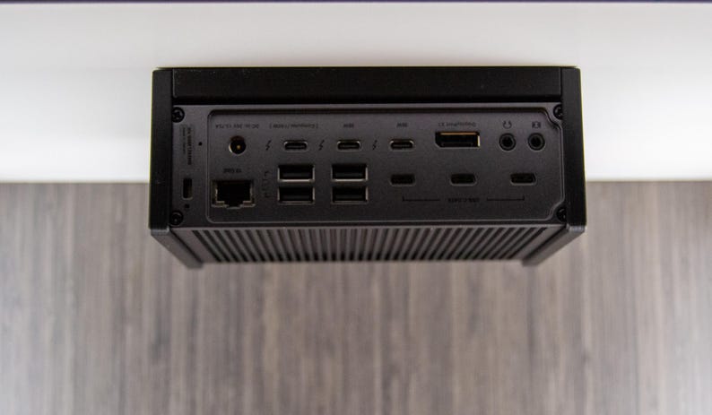 Op de afbeelding: Een zwart elektronisch apparaat met meerdere poorten, waaronder USB, HDMI en Ethernet. Het apparaat heeft een geribbelde onderkant en is onder een wit oppervlak gemonteerd. De poorten zijn gemarkeerd met verschillende symbolen.
