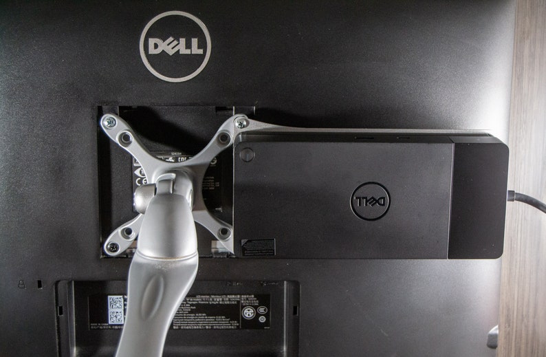May include: A black Dell monitor with a silver adjustable arm attached to the back. A black Dell docking station is attached to the back of the monitor.