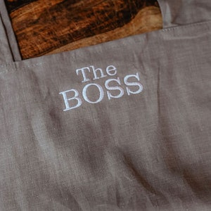 Peut inclure: Un tablier en lin gris avec le texte "The BOSS" brod&eacute; en blanc.