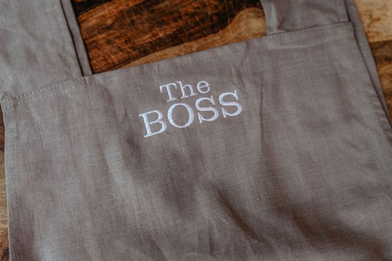 Peut inclure: Un tablier en lin gris avec le texte "The BOSS" brod&eacute; en blanc.