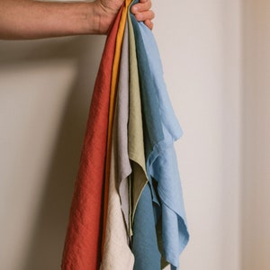 May include: A set of seven linen tea towels in various colors, including red, orange, yellow, green, blue, and gray. The towels are folded and held together in a person's hand.