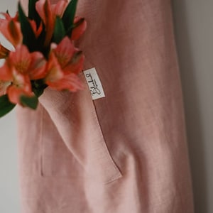 Peut inclure: Un tablier en lin rose poudr&eacute; avec une grande poche et une &eacute;tiquette blanche sur laquelle est inscrit "Blushla" en lettres noires.