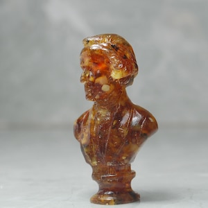 May include: A small amber bust of a person with a detailed face and intricate details throughout the bust.