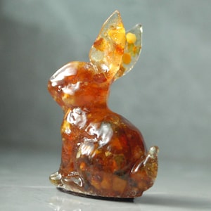 May include: A small, amber-colored rabbit figurine. The sculpture has a glossy finish and is made of a translucent material, with visible inclusions of darker amber and yellow hues. The rabbit is in a seated position with its ears perked up.