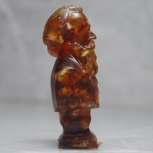May include: A small amber bust of a man with a beard. The bust is made of amber with visible inclusions. The bust is on a white background.