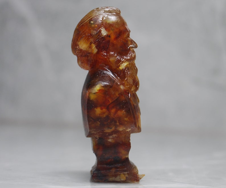 May include: A small amber bust of a man with a beard. The bust is made of amber with visible inclusions. The bust is on a white background.