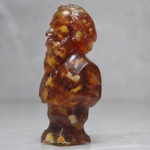 May include: A small amber-colored figurine of a man with a beard and mustache. The figurine is made of a translucent material that allows you to see through it. The figurine is standing on a white surface.