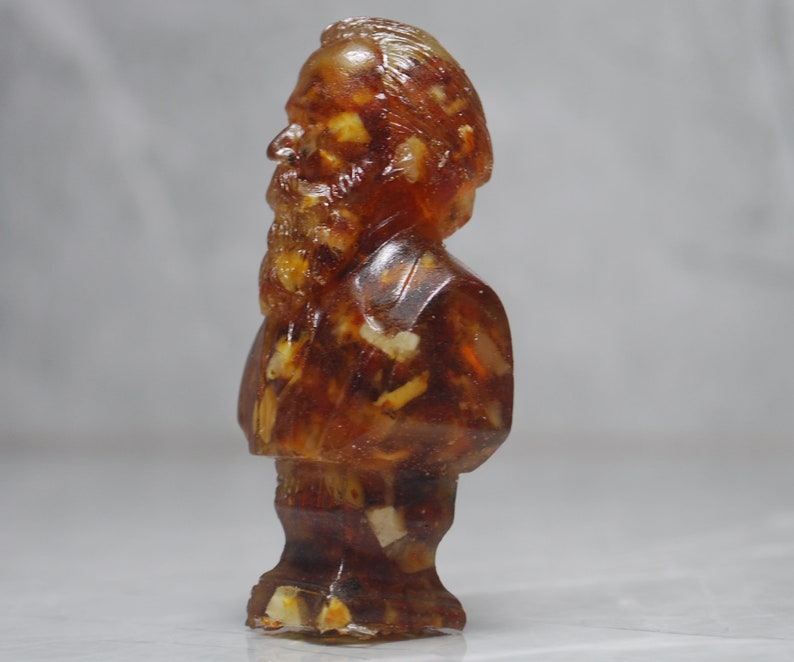 May include: A small amber-colored figurine of a man with a beard and mustache. The figurine is made of a translucent material that allows you to see through it. The figurine is standing on a white surface.