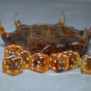 Puede incluir: Un juego de ocho dados de color ámbar con puntos blancos y un soporte para dados de color ámbar a juego con protuberancias en forma de cuerno. Los dados son translúcidos, revelando un interior texturizado. El soporte para dados está en el fondo.