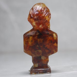 May include: A small amber bust sculpture of a person with a detailed head and torso. The sculpture is made of many small pieces of amber that are fused together.