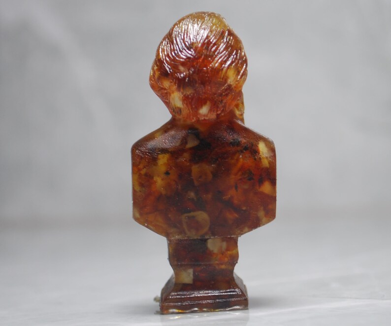 May include: A small amber bust sculpture of a person with a detailed head and torso. The sculpture is made of many small pieces of amber that are fused together.